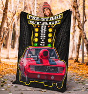 A person outdoors in a wooded, autumn setting holds up a large printed blanket or throw. The blanket features a black background with tire tread graphics, a marquee-style 'PRE-STAGE DRACING RACING' sign with yellow and green lights, and a red race car graphic outlined in yellow.