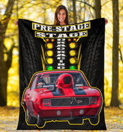 A woman stands outdoors in a wooded area holding up a large black fleece blanket or poster print featuring a red race car and the words 'PRE-STAGE DRAG RACING'.