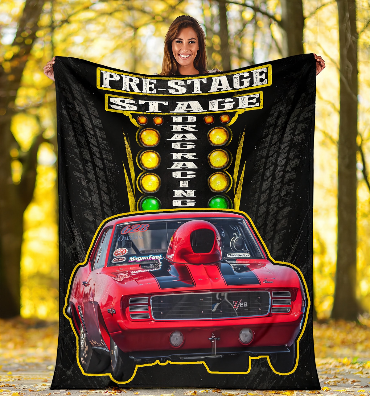A woman stands outdoors in a wooded area holding up a large black fleece blanket or poster print featuring a red race car and the words 'PRE-STAGE DRAG RACING'.