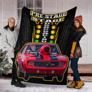 Two people holding up a large black fleece blanket featuring a red muscle car, racing lights, and the text 'PRE-STAGE DRAG RACING' in a snowy outdoor setting in front of a white house.