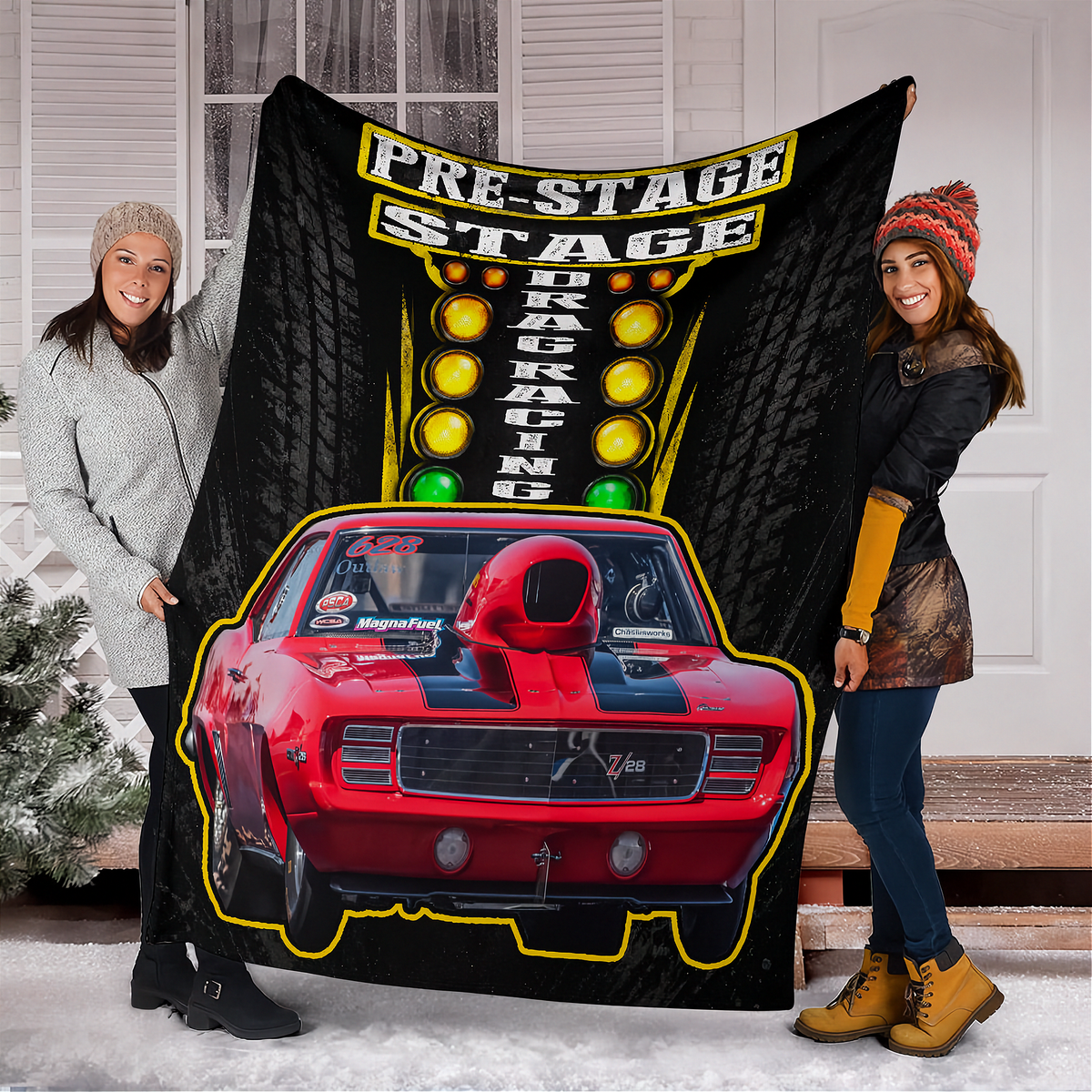 Two people holding up a large black fleece blanket featuring a red muscle car, racing lights, and the text 'PRE-STAGE DRAG RACING' in a snowy outdoor setting in front of a white house.