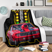 A fleece throw blanket draped over a gray sofa in a living room setting, featuring a graphic print of a red muscle car with a drag racing light panel above it.