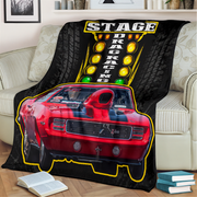 A black throw blanket draped over a beige sofa in a living room, printed with a red drag racing car and a large 'STAGE DRAG RACING' graphic with yellow lights and tire tracks.