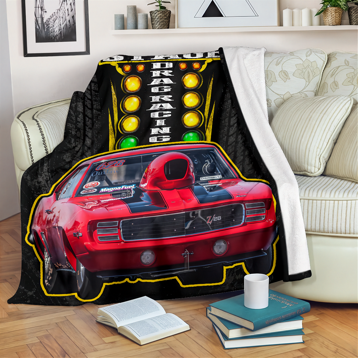 A black throw blanket draped over a cream sofa in a living room, featuring a printed design of a red muscle car in front of a yellow racing light sign with the words 'DRAG RACING'.