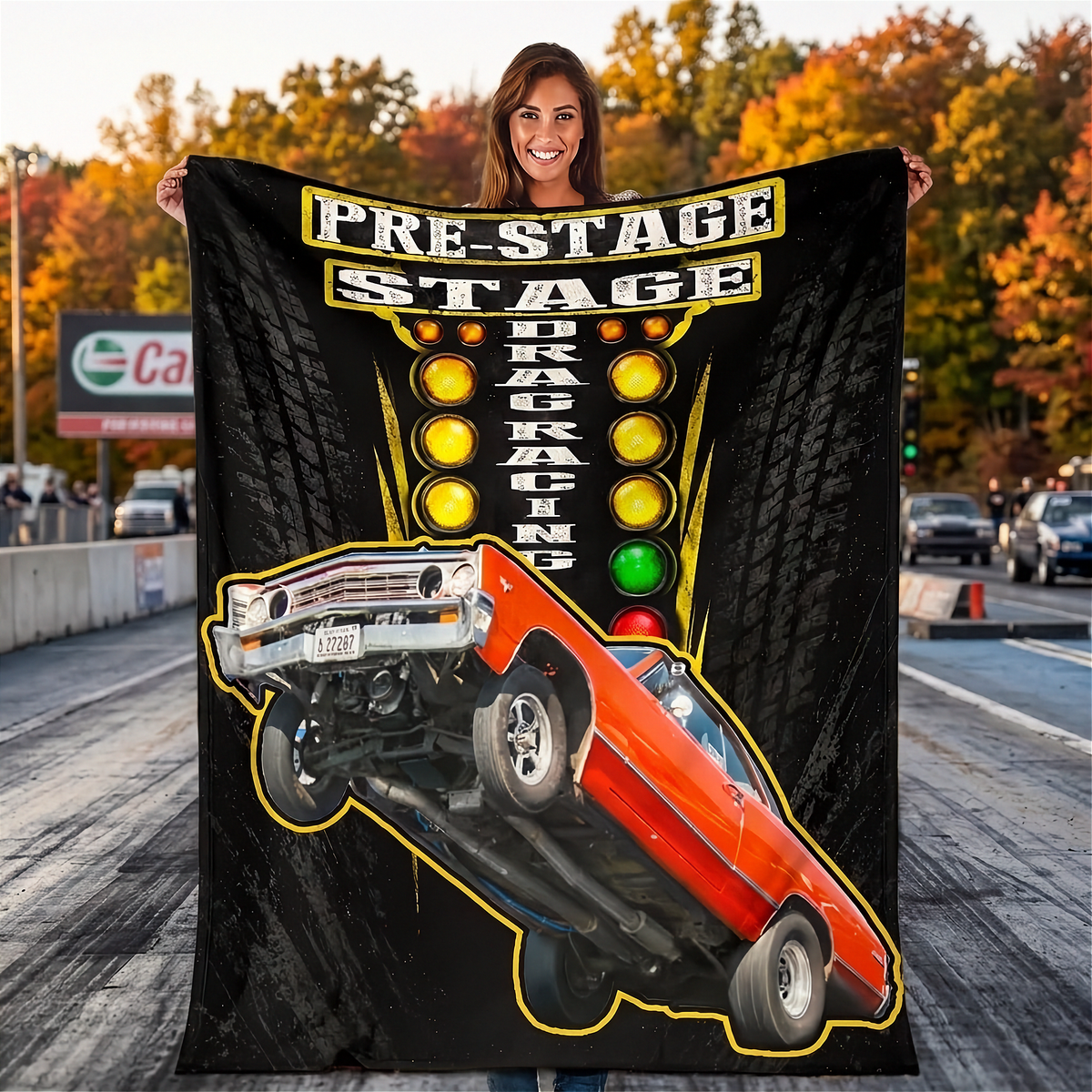 A woman stands on a road holding up a large black fleece blanket featuring a graphic of an orange muscle car doing a wheelie with the text 'PRE-STAGE DRAG RACING' and illuminated race lights.