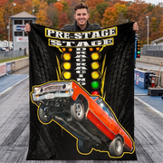 A man stands on a drag strip holding up a large black fleece blanket or poster print featuring a red muscle car doing a wheelie, with the words 'PRE-STAGE DRAG RACING' and yellow light bulbs on a tire-pattern background.