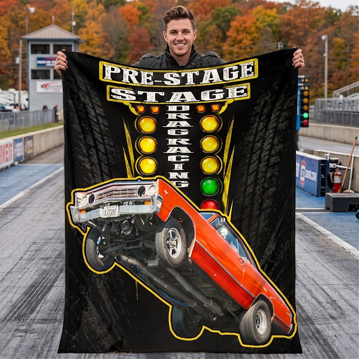 A man stands on a drag strip holding up a large black fleece blanket or poster print featuring a red muscle car doing a wheelie, with the words 'PRE-STAGE DRAG RACING' and yellow light bulbs on a tire-pattern background.