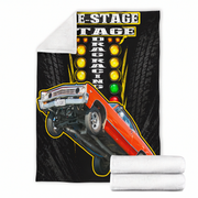 A fleece throw blanket featuring a vintage drag racing graphic with an orange car jumping over a track light signal, shown flat with a folded corner revealing a white sherpa lining. Two rolled white sherpa blankets are stacked at the bottom right on a white background.