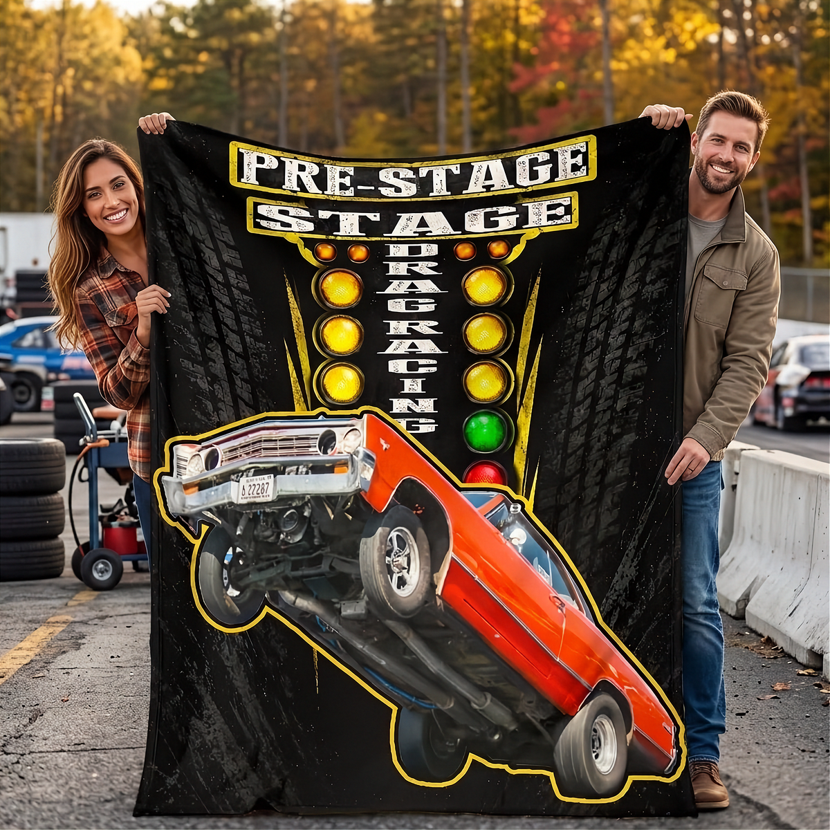 A man and a woman hold up a large black fleece blanket featuring a graphic of an orange muscle car doing a donut on track, with 'PRE-STAGE DRAG RACING' and illuminated stoplight graphics. The photo is taken outdoors on a racetrack with trees in the background.