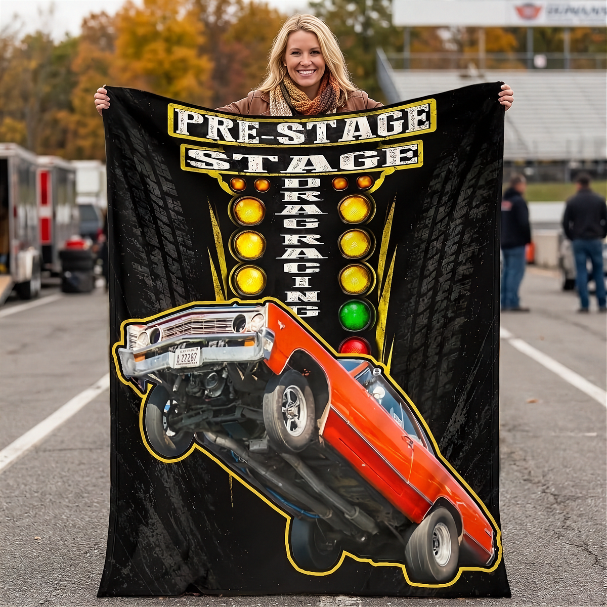 A woman stands on an outdoor racetrack holding up a large black fleece blanket or throw. The blanket features a graphic of an orange muscle car flipping over, yellow and red lights, and the words 'PRE-STAGE DRAG RACING'.