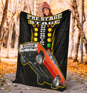 A woman outdoors in a wooded path holds up a large black blanket or throw featuring a racing graphic with the words 'PRE-STAGE DRAG RACING,' a red race car, and yellow traffic-light-style marquee lights.