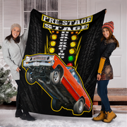 A black fleece blanket featuring a 'Pre-Stage Drag Racing' graphic with an orange drag car and yellow tire tracks, held up by two smiling women in front of a white house door.