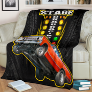 A black throw blanket featuring a graphic of a red drag racing car on its rear wheels with a yellow-tiled track sign and the words 'STAGE DRAG RACING', displayed draped over a light beige sofa in a living room setting.