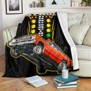 A black throw blanket draped over a cream sofa in a living room setting, featuring a graphic print of an orange drag racing car on a tilted track with yellow lights and the words 'DRAG RACING'.