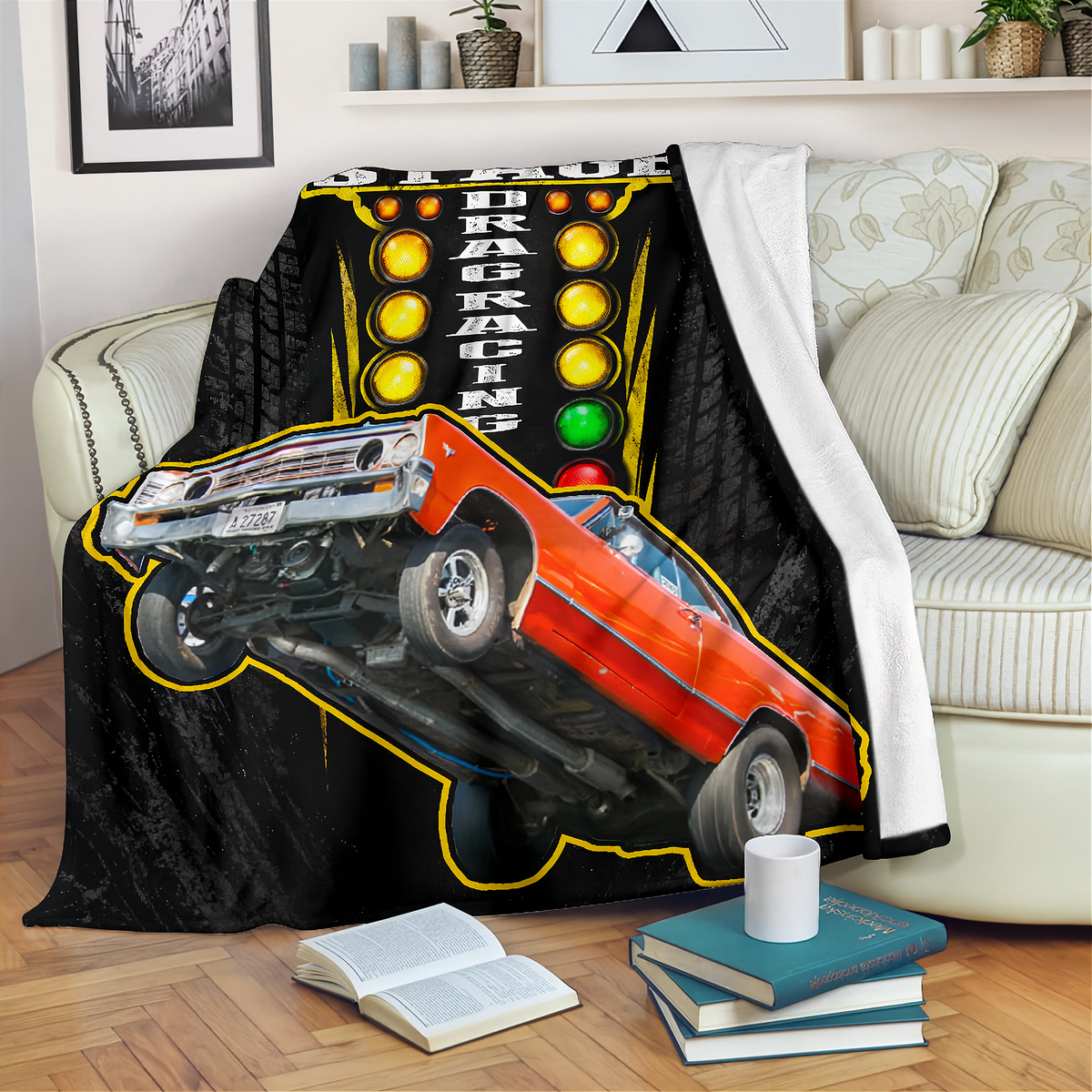 A black throw blanket draped over a cream sofa in a living room setting, featuring a graphic print of an orange drag racing car on a tilted track with yellow lights and the words 'DRAG RACING'.