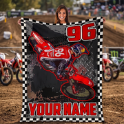A woman stands on a dirt track holding up a large customizable motocross blanket. The blanket features a graphic of a red dirt bike with the number 96, a checkerboard border, and the text 'YOUR NAME'.