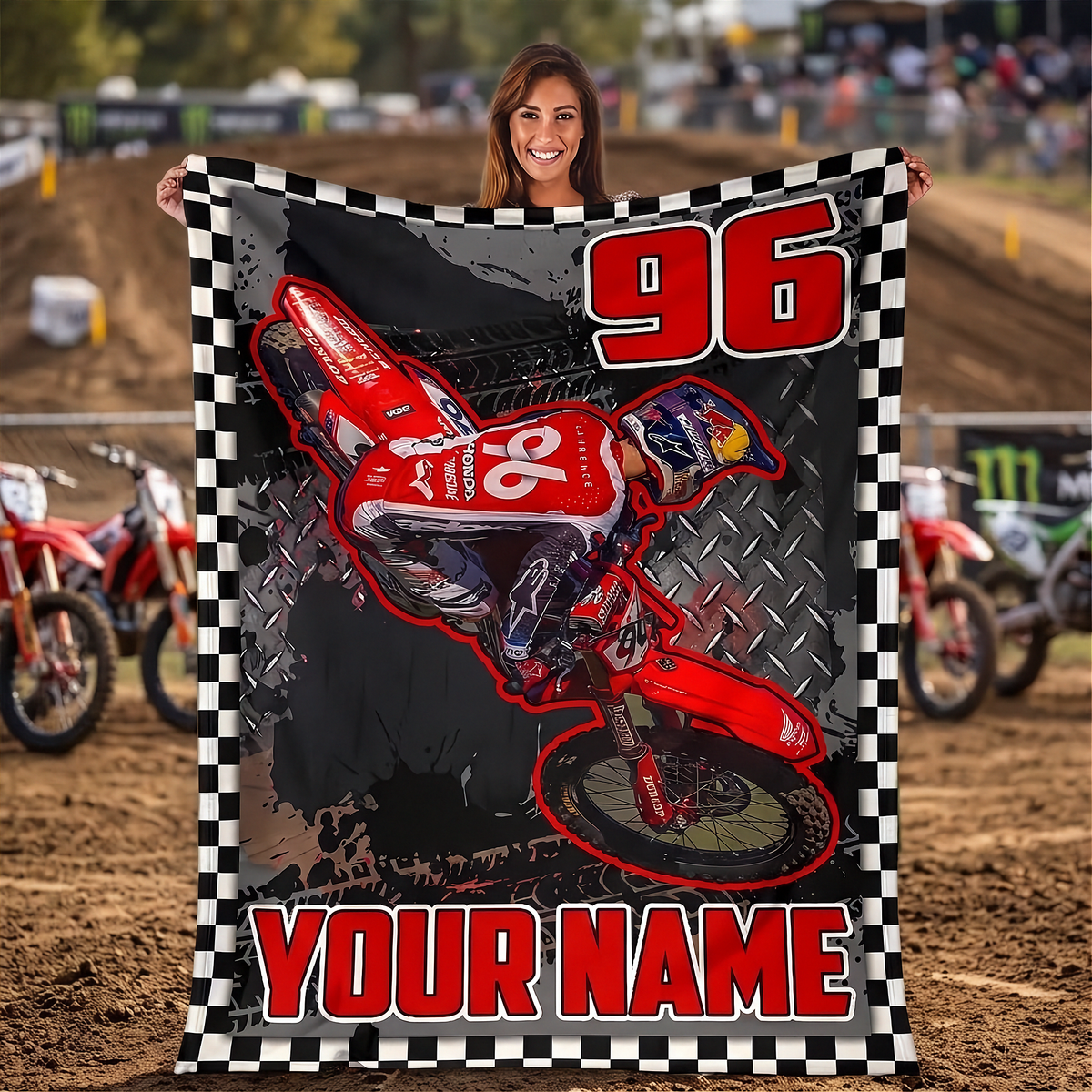 A woman stands on a dirt track holding up a large customizable motocross blanket. The blanket features a graphic of a red dirt bike with the number 96, a checkerboard border, and the text 'YOUR NAME'.