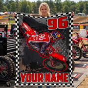 A woman holds up a large custom motocross blanket featuring a red dirt bike graphic, the number 96, and 'YOUR NAME' in red text, with a black-and-white checkered border. A red motocross bike and pit area equipment are visible in the outdoor background.