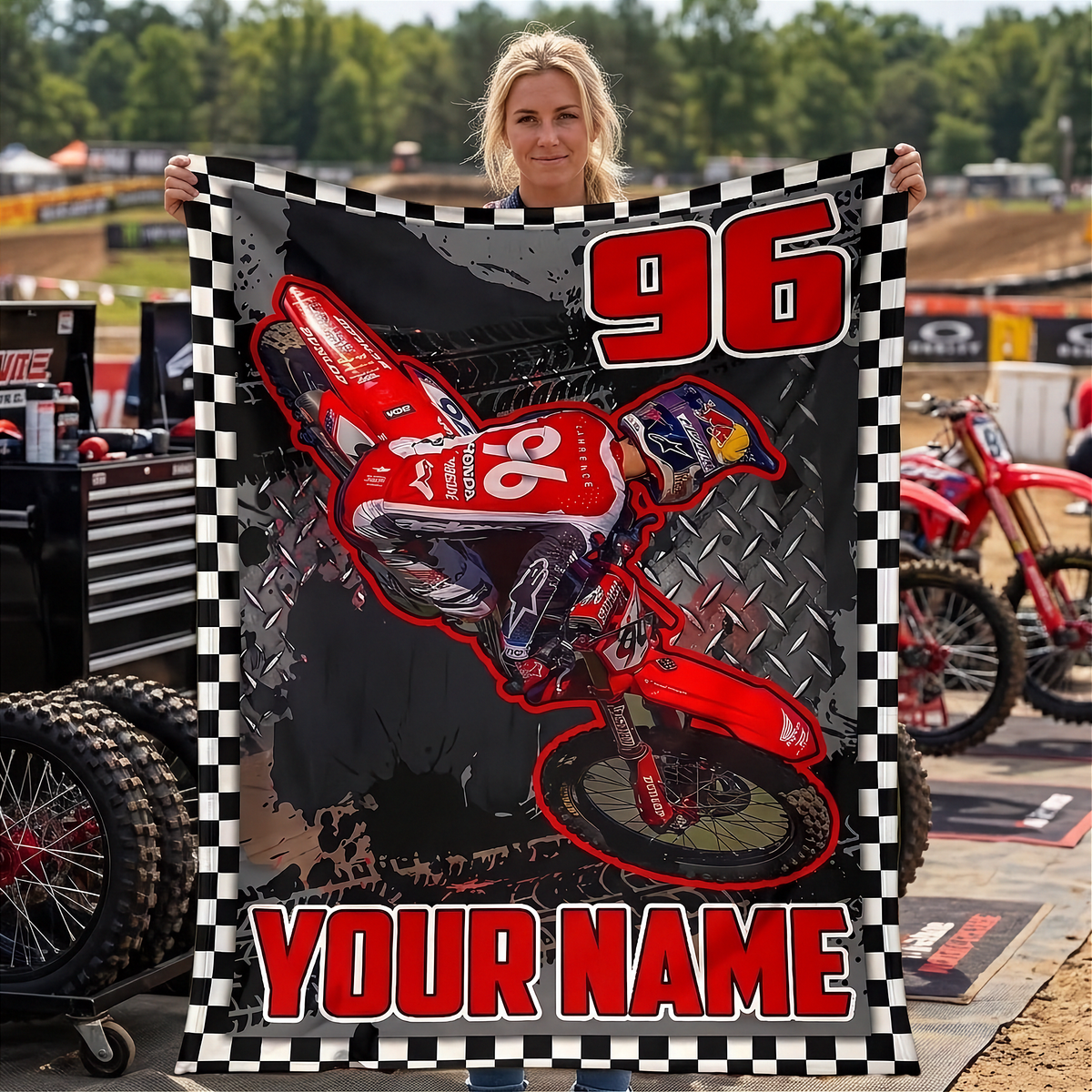 A woman holds up a large custom motocross blanket featuring a red dirt bike graphic, the number 96, and 'YOUR NAME' in red text, with a black-and-white checkered border. A red motocross bike and pit area equipment are visible in the outdoor background.