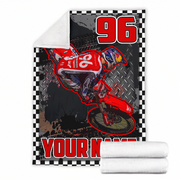 A custom printed fleece blanket featuring a red dirt bike racer graphic with the number 96, a black-and-white checkered border, and a white sherpa lining shown on the folded corner. Two rolled white blankets are stacked in front on a plain white background.