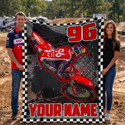A man and a woman stand outdoors holding a large custom motocross blanket. The blanket features a red dirt bike graphic, the number 96, and the text 'YOUR NAME' on a black-and-white checkered border.