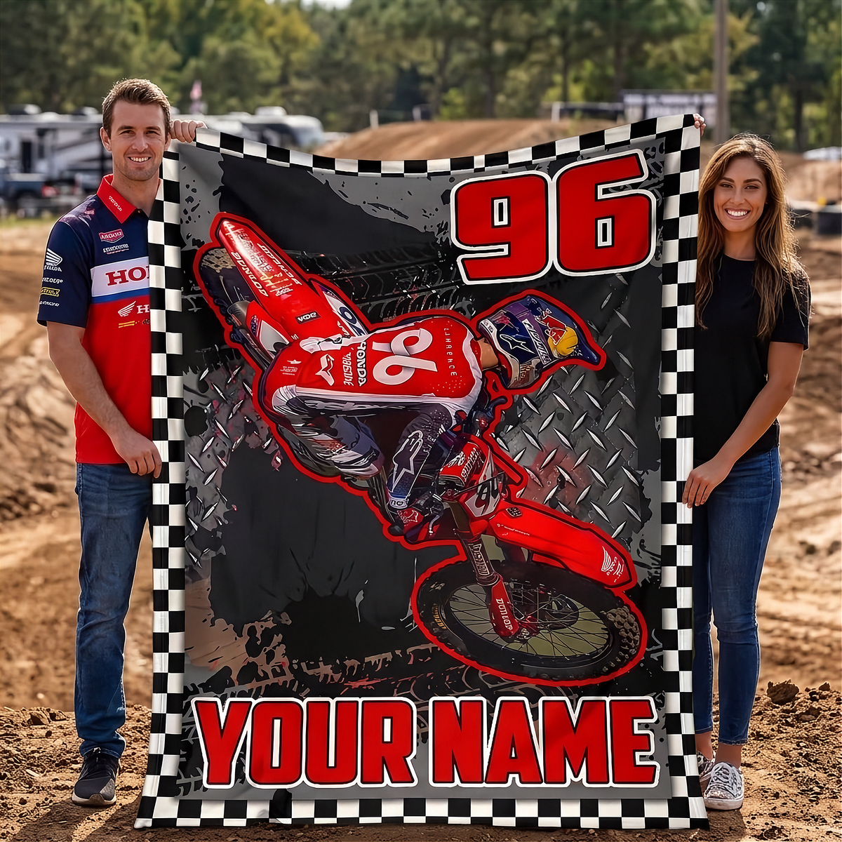 A man and a woman stand outdoors holding a large custom motocross blanket. The blanket features a red dirt bike graphic, the number 96, and the text 'YOUR NAME' on a black-and-white checkered border.