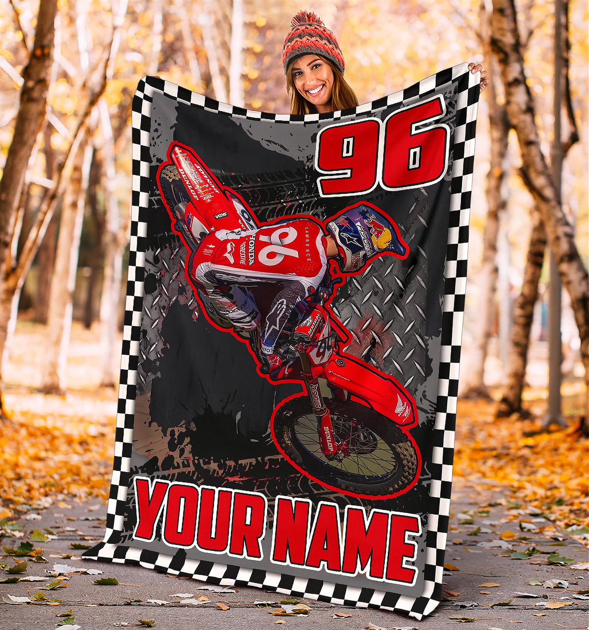 A person wearing a red beanie holds up a large custom-printed blanket featuring a red dirt bike, the number 96, and 'YOUR NAME' text, set against an outdoor autumn forest background.