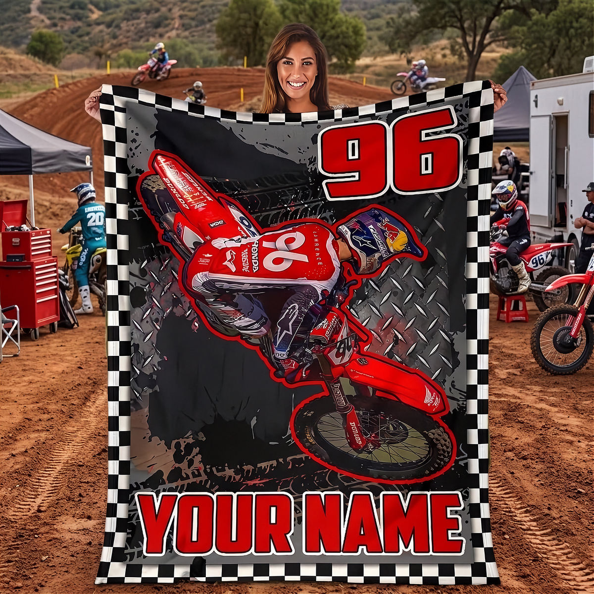 A custom motocross-themed blanket held up by a woman at an outdoor dirt track event. The blanket features a red and white dirt bike graphic, the number 96, and 'YOUR NAME' in red text on a black and white checkered border.