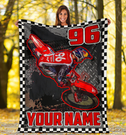A woman outdoors in a forest holding up a large custom-printed blanket. The blanket features a red dirt bike graphic, the number 96, and 'YOUR NAME' in red text, framed by a black and white checkered border.