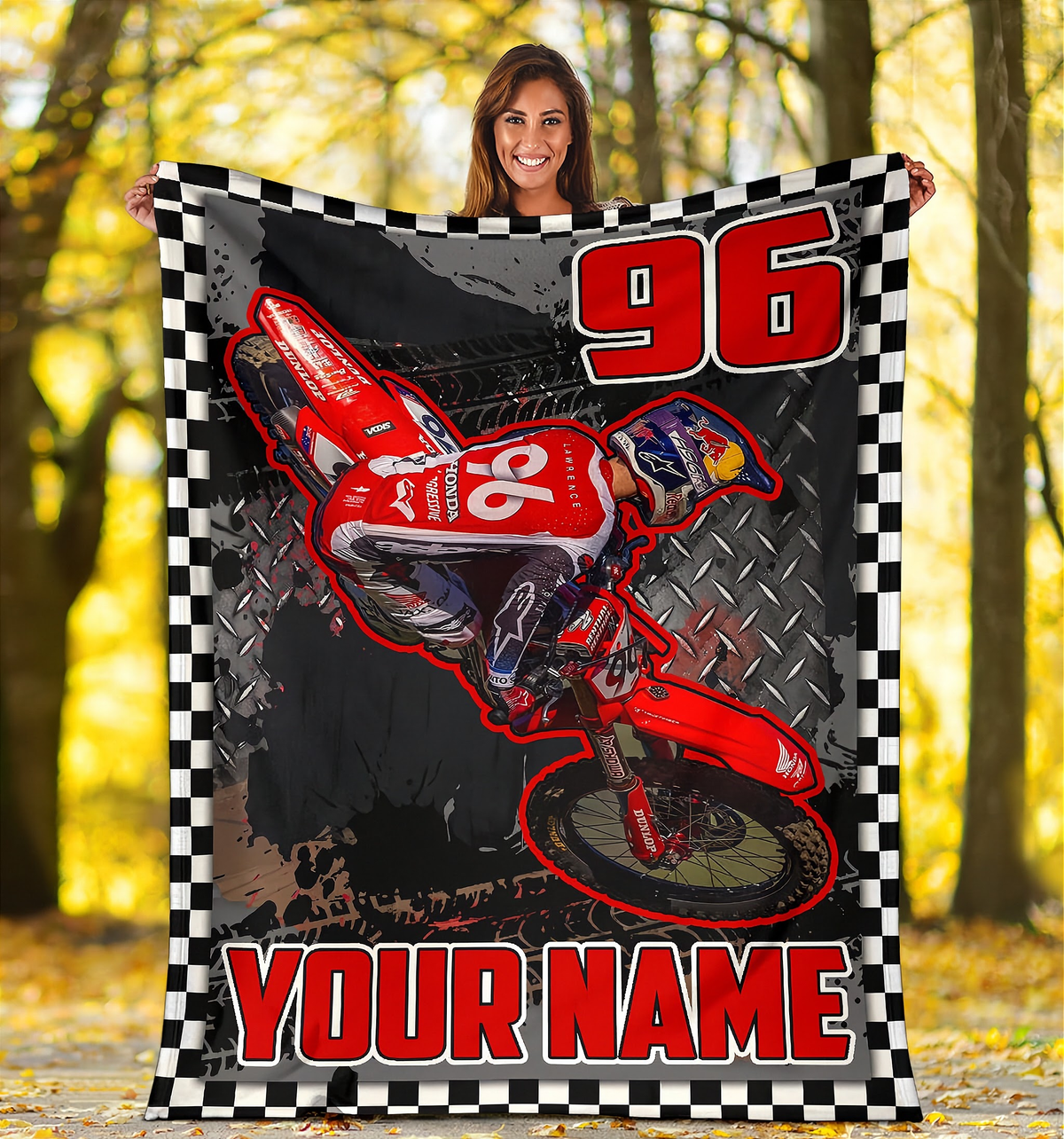 A woman outdoors in a forest holding up a large custom-printed blanket. The blanket features a red dirt bike graphic, the number 96, and 'YOUR NAME' in red text, framed by a black and white checkered border.
