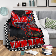 A custom printed motorcycle blanket draped over a gray sofa in a living room setting. The blanket features a red dirt bike graphic, a black and white checkered border, and the text 'YOUR NAME' in bold red letters.