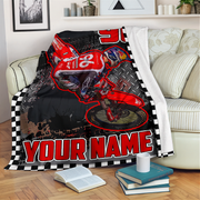 A custom printed blanket draped over a sofa in a living room setting. The blanket features a red dirt bike racer graphic on a black and white checkered background with the text 'YOUR NAME' in bold red letters.