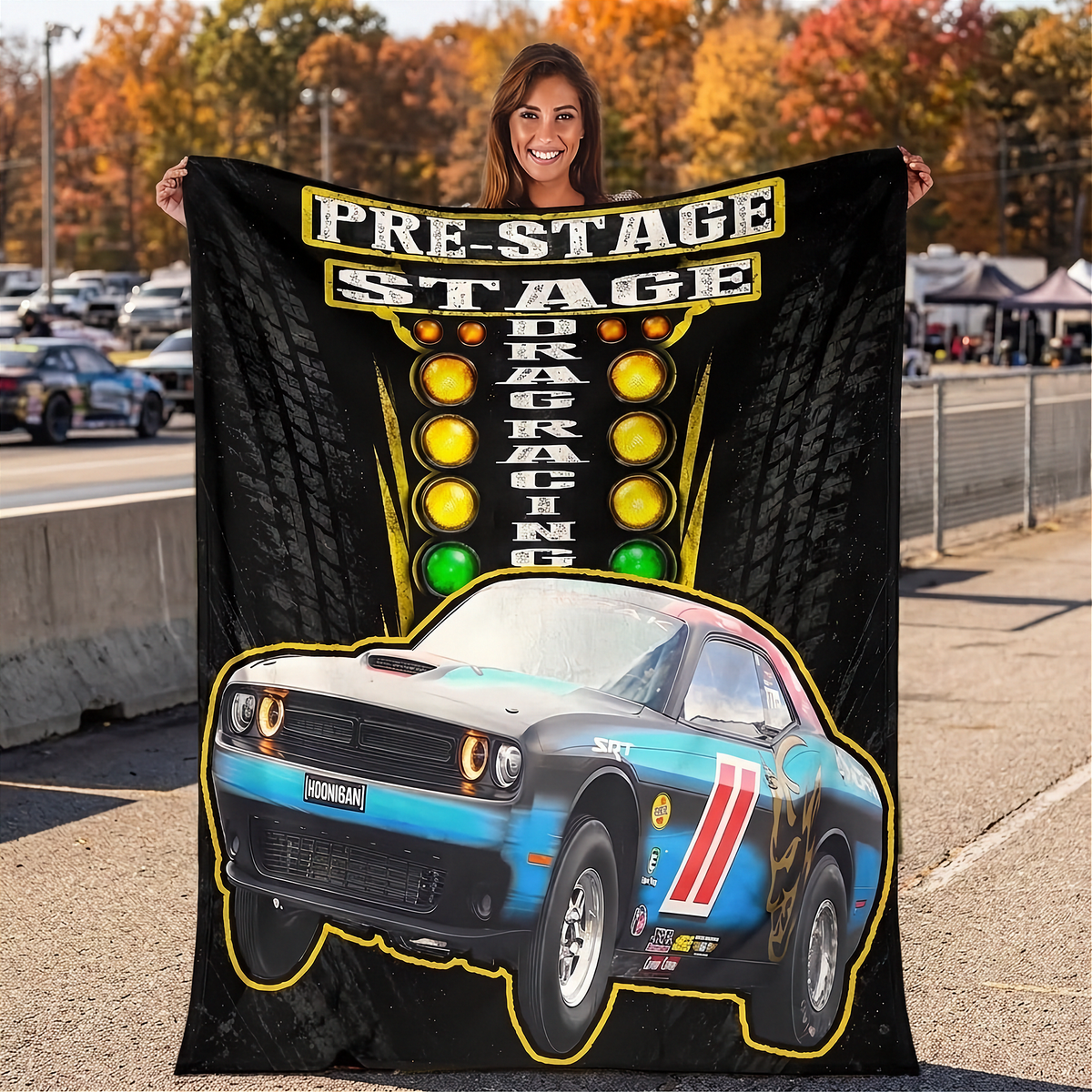 A woman stands outdoors holding a large black printed blanket or poster featuring a blue and black race car, yellow 'PRE-STAGE DRAG RACING' text, and illuminated light graphics. The background shows a racetrack scene with trees and cars.