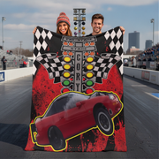 A couple stands on a racetrack holding up a large custom fleece blanket featuring a red race car, checkered flags, and a pre-stage track graphic.