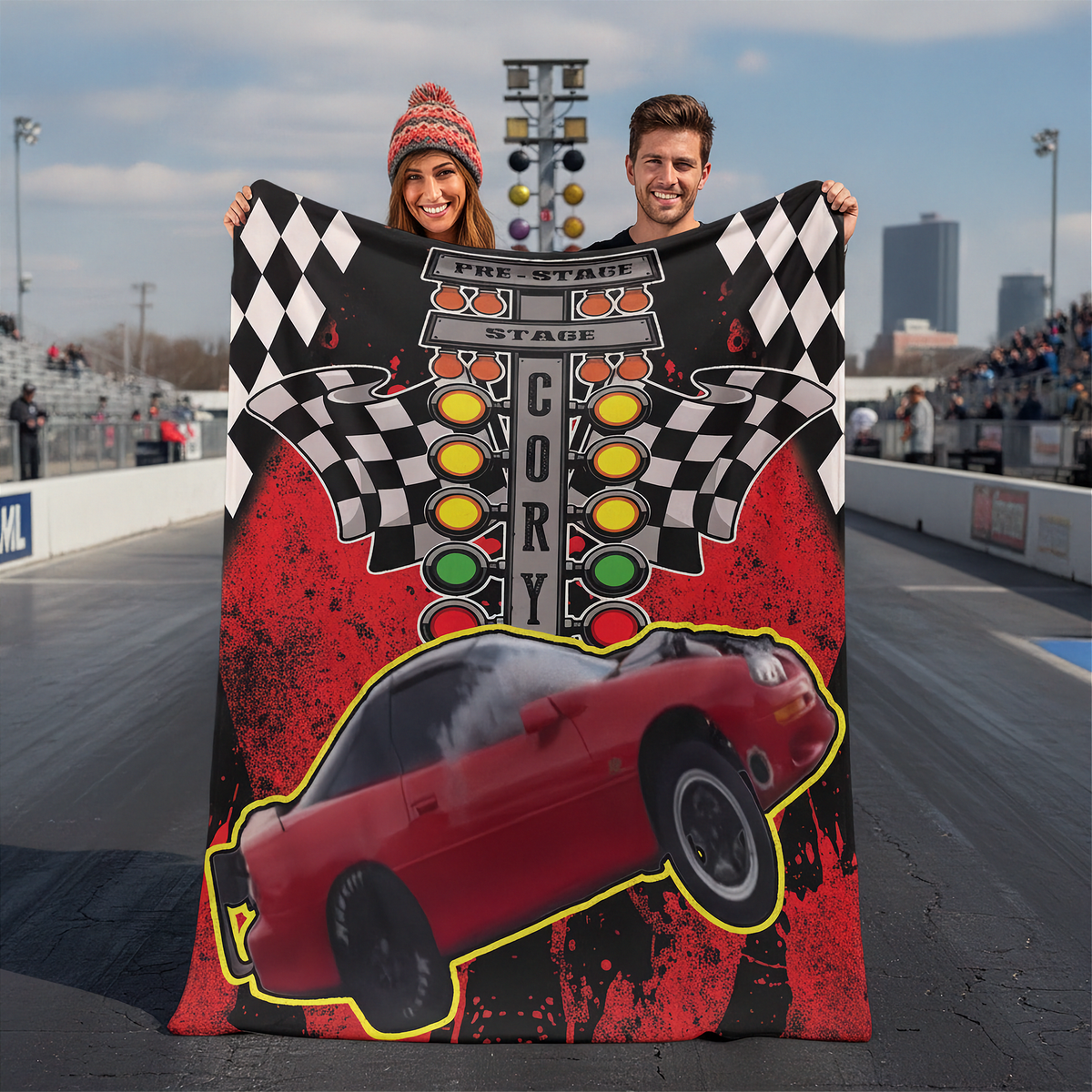 A couple stands on a racetrack holding up a large custom fleece blanket featuring a red race car, checkered flags, and a pre-stage track graphic.