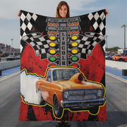 A woman stands on a racetrack holding up a large printed blanket or tapestry featuring a vintage orange race car, checkered flags, and the words 'PRE STAGE LINCOLN'.