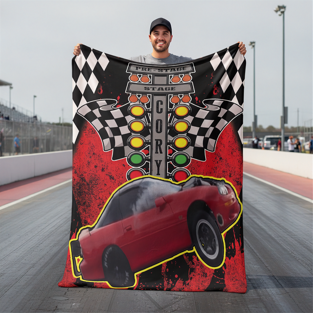 A man standing on a racetrack holds up a large printed blanket or throw featuring a red race car, checkered flags, a starting line graphic, and the word 'CORY' on a red, black, and gray background.