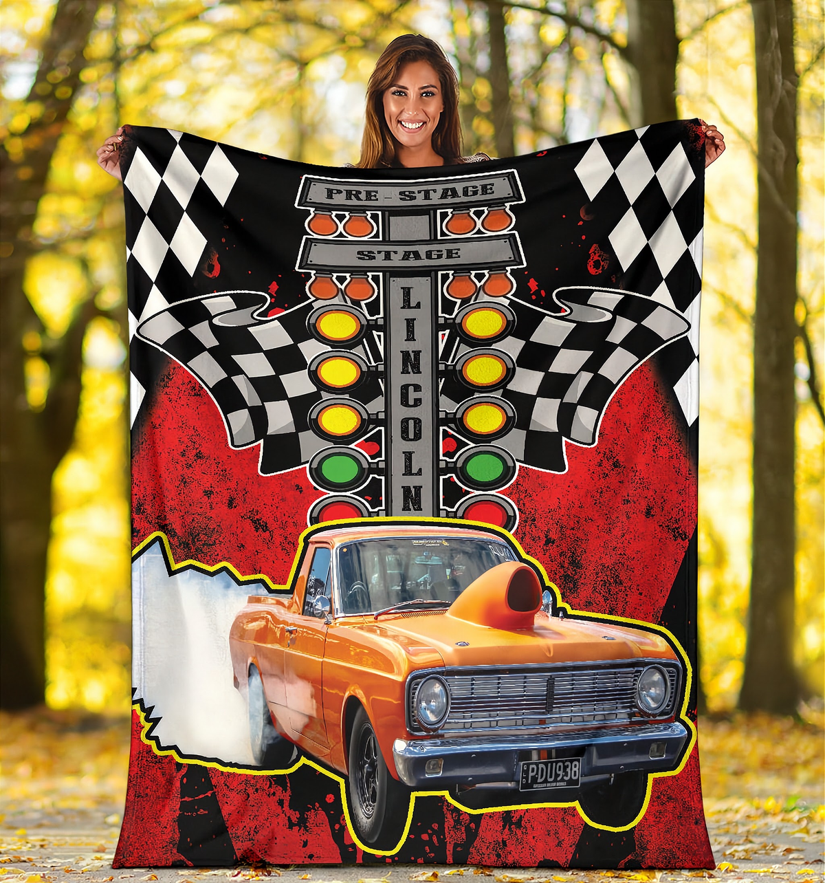 A woman holds up a large printed blanket or tapestry outdoors in a wooded area. The blanket features a racing design with a black-and-white checkered flag, a vertical 'LINCOLN' sign, yellow and green traffic lights, red distressed background areas, and an orange race car with a flame effect.