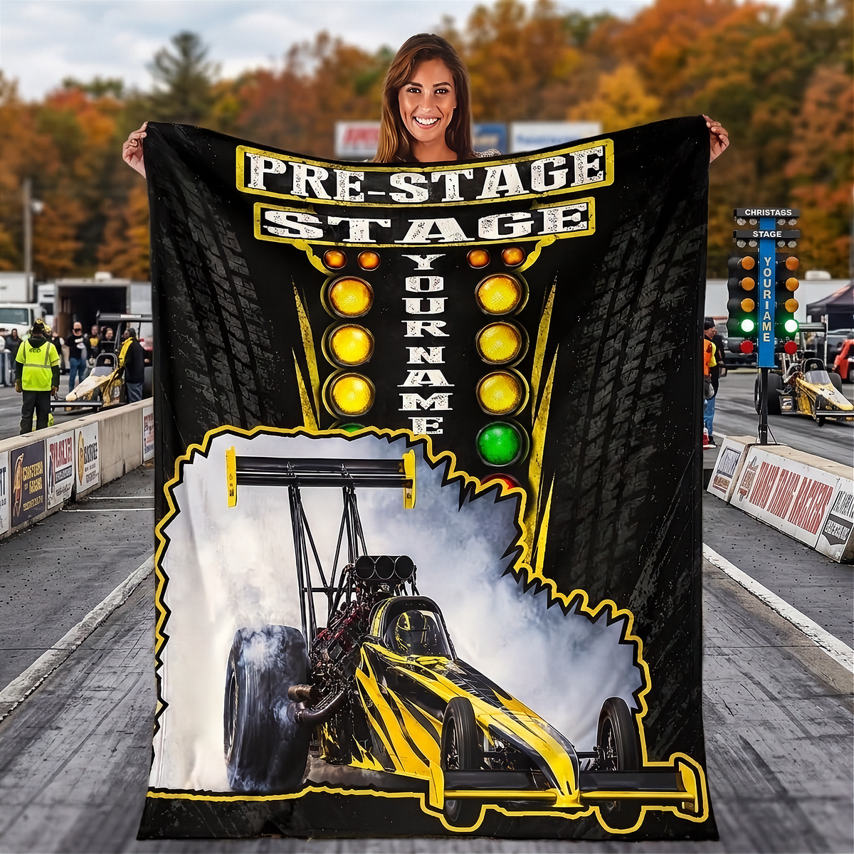 A woman stands on a racetrack holding up a large custom fleece blanket. The blanket features a black background with yellow and white text reading 'PRE-STAGE STAGE YOUR NAME', a yellow and black drag racer graphic, and a white smoke effect.