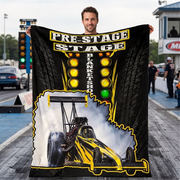 A man stands on a racetrack holding up a large black printed blanket or poster featuring a yellow drag racer, tire tracks, and the words 'PRE-STAGE BLANKET SHOT'.