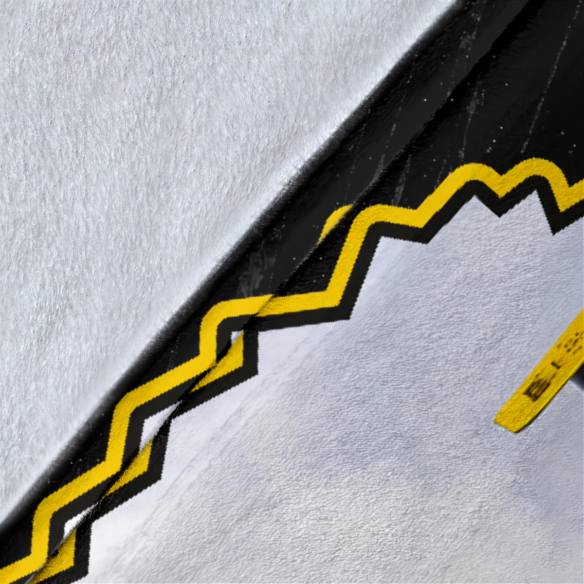 A close-up of a textured white surface with a bold black and yellow zigzag graphic running diagonally across the frame, plus a small yellow tag in the lower right corner.
