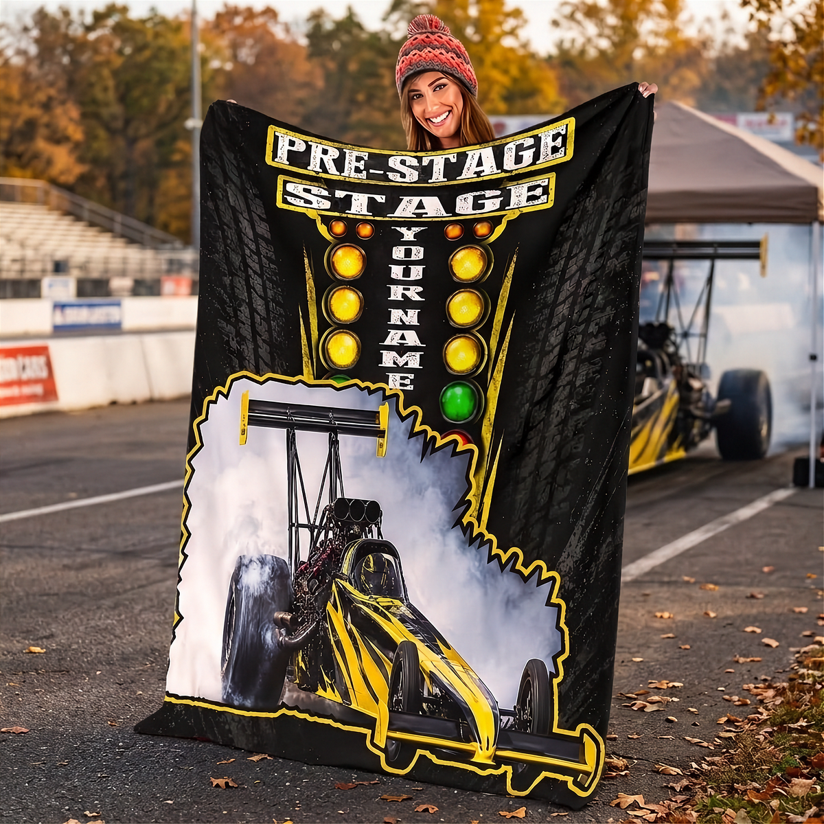 A woman outdoors on a racetrack holds up a large custom-printed black blanket or throw featuring a yellow and black race car, smoke graphic, and the words 'PRE-STAGE YOUR NAME'. A race car is visible in the background near a canopy tent.