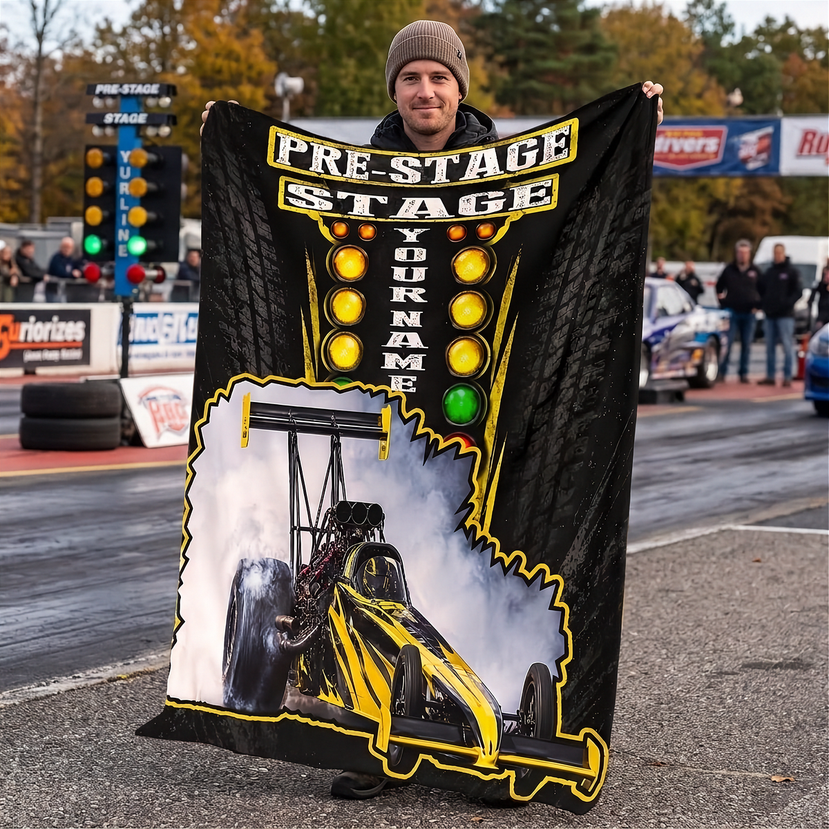 A man stands on a racetrack holding up a large custom-printed blanket or throw. The blanket features a black background with yellow and white tire tread graphics, a yellow race car, and the text 'PRE-STAGE STAGE' and 'YOUR NAME'.