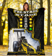 A woman stands outdoors in a wooded area holding up a large custom-printed blanket or throw. The blanket features a black background with tire tread graphics, yellow and white racing-style text reading 'PRE-STAGE STAGE' and 'YOUR NAME', a green light, and a yellow-and-black race car with smoke.