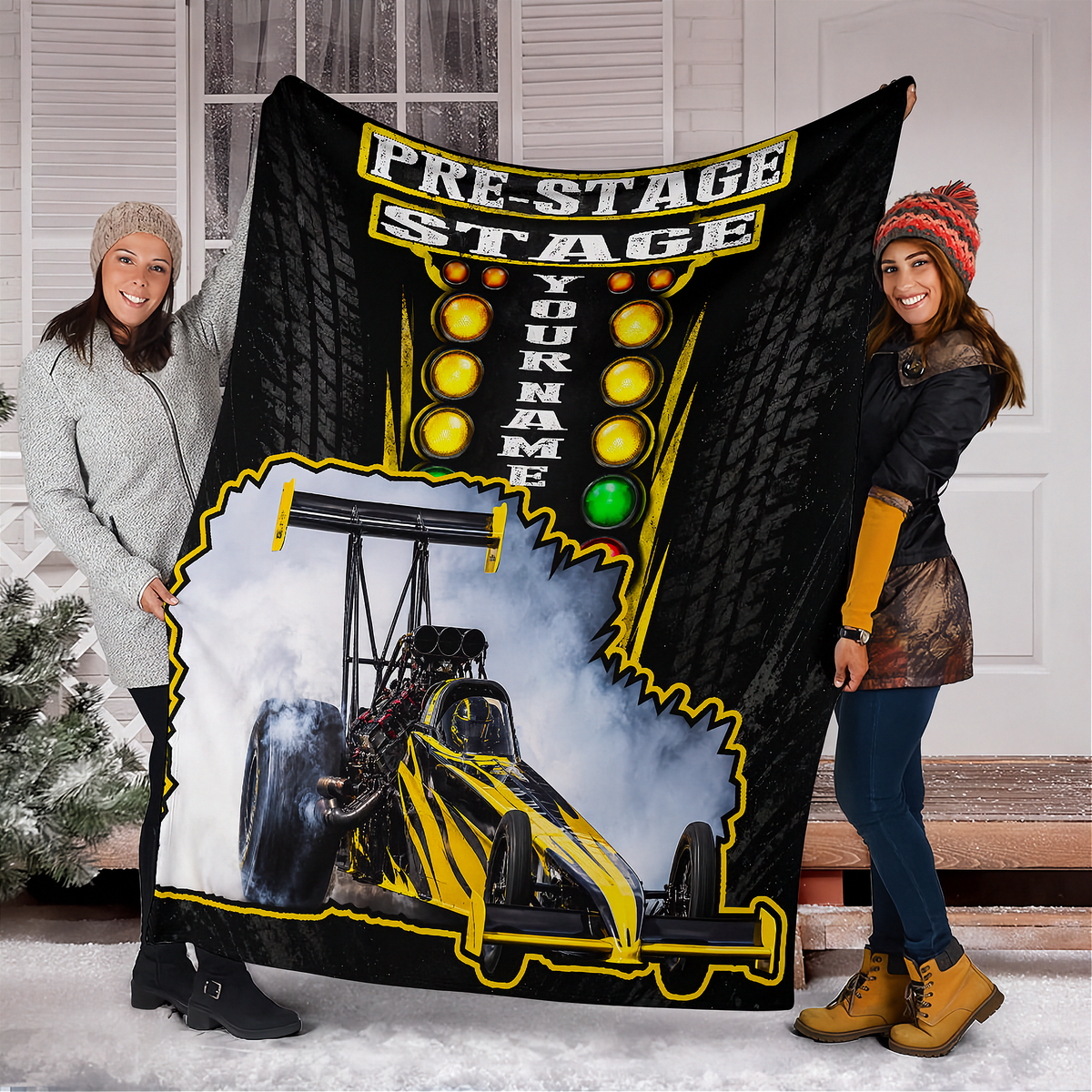 Two people holding up a custom fleece blanket featuring a drag racer graphic and 'PRE-STAGE STAGE YOUR NAME' text, set against a snowy porch background.