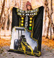 A woman wearing a red knit beanie holds up a large custom-printed blanket outdoors in a wooded area with fallen leaves. The blanket is black with yellow and white graphics, featuring the text 'PRE-STAGE STAGE', 'YOUR NAME', a traffic light motif, and a yellow drag racer illustration.