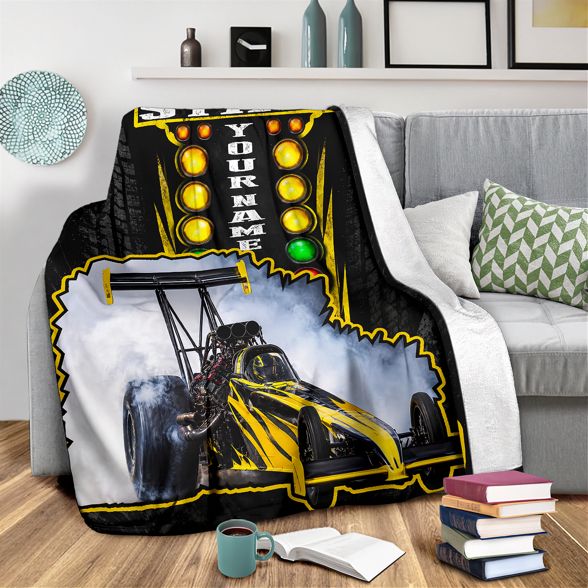 A custom race car blanket draped over a gray sofa in a living room setting. The blanket features a black and yellow drag racing design with a customizable name print area, set against a background of a gray couch, wooden floor, and decorative pillows.