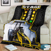 A custom printed fleece blanket draped over a light beige sofa in a living room setting. The blanket features a black and yellow drag racing theme with a graphic of a race car smoking tires, a yellow 'STAGE YOUR NAME' light bar design, and a white smoke border.