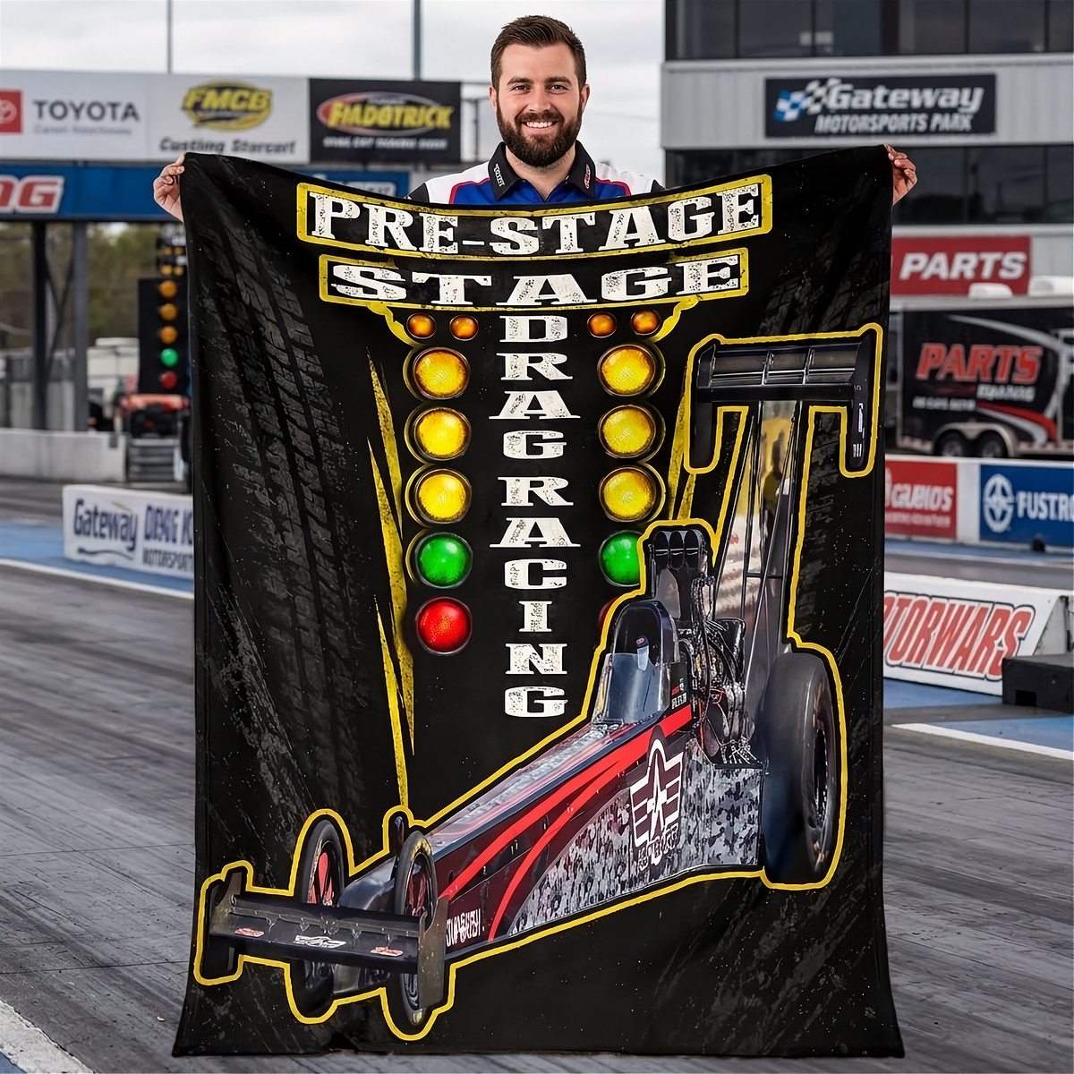 A man stands on a drag racing track holding up a large black fleece blanket or throw featuring a drag racer car, tire patterns, and the words 'PRE-STAGE DRAG RACING' with illuminated light bulbs.