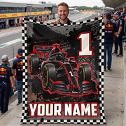 A man standing on a racetrack holds up a large customizable blanket or tapestry featuring a black and red Formula 1 race car, a large number 1, and the text 'YOUR NAME' on a checkered border. Other people in Red Bull team apparel are visible in the background on the track.